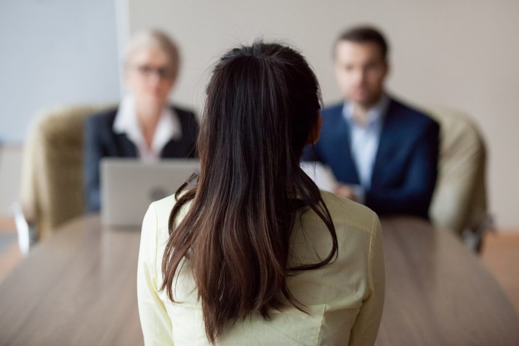 Businesswoman and businessman HR manager interviewing woman. Candidate female sitting her back to camera, focus on her, close up rear view, interviewers on background. Human resources, hiring concept